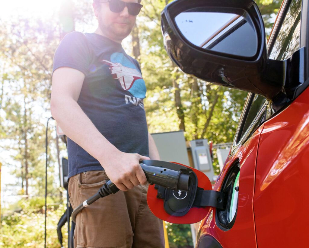 A low angle shot of a man wearing a blue shirt and sunglasses plugging in a red electric vehicle at a charging station.